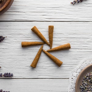 Op de afbeelding: Een set van zes bruine wierookkegels, gerangschikt in een stervorm op een wit houten oppervlak. Gedroogde lavendelbloemen zijn rond de kegels verspreid.