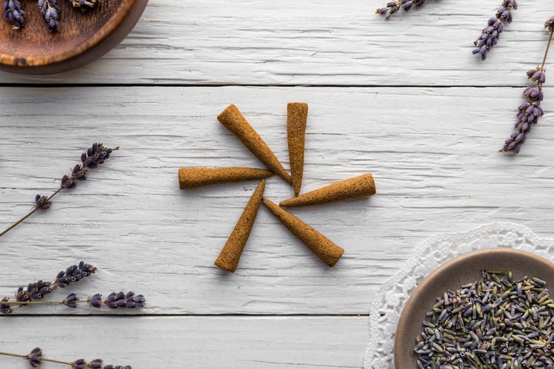 Op de afbeelding: Een set van zes bruine wierookkegels, gerangschikt in een stervorm op een wit houten oppervlak. Gedroogde lavendelbloemen zijn rond de kegels verspreid.