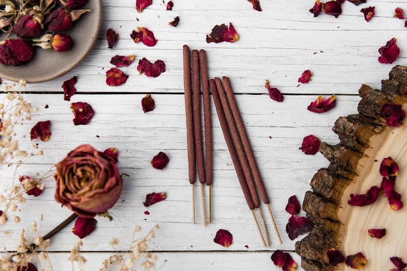 Peut inclure: Cinq b&acirc;tonnets d'encens bruns avec une base en bois naturel, dispos&eacute;s sur une surface en bois blanc avec des p&eacute;tales de rose s&eacute;ch&eacute;s &eacute;parpill&eacute;s autour.