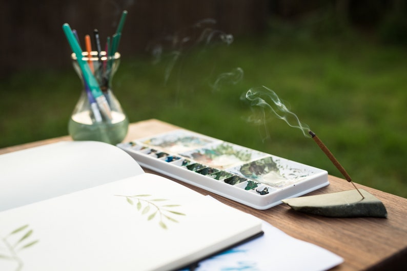 A white plastic watercolor palette with various paint pigments, a glass jar containing several paintbrushes, an open sketchbook with a green leaf illustration, and a burning incense stick held by a dark stone holder.