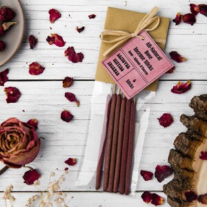 Peut inclure: Un paquet de six b&acirc;tonnets d'encens &agrave; la rose enti&egrave;rement naturels dans un sac en plastique transparent avec une &eacute;tiquette rose et blanche qui indique "All-Natural Rose Incense Sticks" et "6 Sticks". Le paquet est entour&eacute; de p&eacute;tales de rose s&eacute;ch&eacute;s sur une surface en bois blanc.