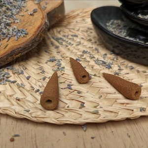 May include: Three brown, cone-shaped incense cones with a hollow centre hole at the base. The incense cones are positioned on a woven straw mat alongside scattered dried lavender buds and a natural wood slice with more lavender.