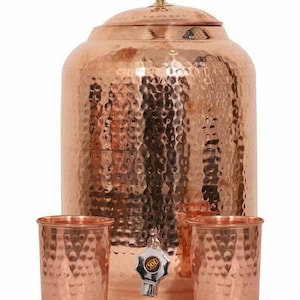 Könnte beinhalten: Ein Kupfer-Wasserspender mit gehämmertem Muster, Deckel und Zapfhahn. Zwei passende Kupferbecher stehen vor dem Spender. Der Spender und die Becher haben eine glänzende, metallische Oberfläche.