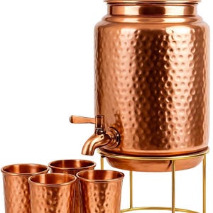Könnte beinhalten: Ein Kupfer-Wasserspender mit gehämmertem Muster, einem Wasserhahn und einem Deckel. Er steht auf einem goldfarbenen Ständer, zusammen mit vier passenden Kupferbechern. Der Spender ist zum Servieren von Getränken bestimmt.
