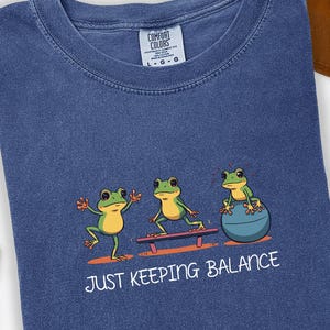 May include: Denim blue t-shirt with a graphic of three cartoon frogs balancing. One frog is on a bench, another on a balance ball, and the third is standing. The text "JUST KEEPING BALANCE" is below the frogs.