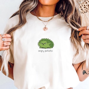 May include: Ivory t-shirt with a green cartoon hedgehog and the words "angry patato." The shirt is worn with light blue denim shorts and a brown belt. The outfit has a casual style. The image also shows a decorative buckle.
