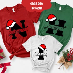 May include: Three short-sleeved t-shirts in red, green, and white, each featuring a black initial with a Santa hat. The red shirt has the name "Nick", the green shirt has "Holly", and the white shirt has "Ivy".