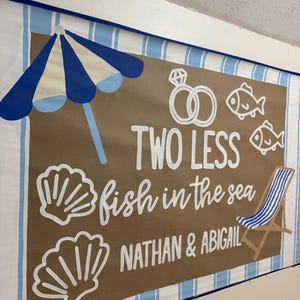 Pode incluir: Uma placa com tema de praia com listras azuis e brancas, um guarda-sol azul e branco e o texto "TWO LESS fish in the sea". Inclui uma cadeira de praia, dois peixes, dois anéis, duas conchas e os nomes "NATHAN & ABIGAIL".