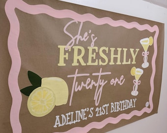 Könnte beinhalten: Braunes Banner mit rosa und gelbem Text: "She's FRESHLY twenty one" und "Adeline's 21st Birthday". Das Banner hat einen rosa Rand, Zitronenillustrationen und Cocktailgrafiken.