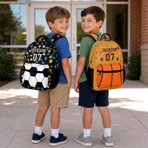 May include: Two backpacks featuring personalized text JACKSON and the number 07. The left backpack is black with a white soccer ball pattern. The right backpack is orange with a basketball design. Both have small sports-themed icons printed on them.