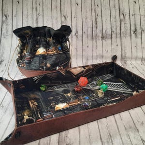May include: A brown leather dice tray with a fantasy-themed print featuring a library interior. The tray has a matching brown leather dice bag with a drawstring closure. The tray is filled with colorful dice.