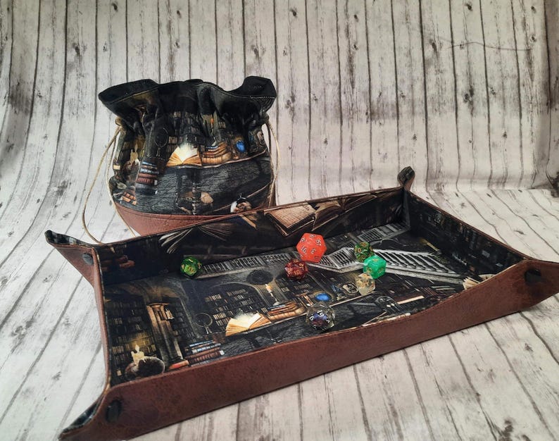 May include: A brown leather dice tray with a fantasy-themed print featuring a library interior. The tray has a matching brown leather dice bag with a drawstring closure. The tray is filled with colorful dice.
