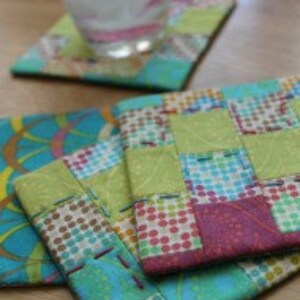 Puede incluir: Cuatro posavasos de tela hechos a mano en una variedad de patrones y colores, incluyendo verde, azul, rosa y marr&oacute;n. Los posavasos est&aacute;n hechos de cuadrados de tela patchwork y tienen un dise&ntilde;o acolchado.