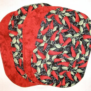 Puede incluir: Juego de cuatro manteles individuales ovalados de color rojo y negro con un patr&oacute;n de chiles. Los manteles individuales tienen un borde rojo y un fondo negro con chiles rojos y hojas verdes.