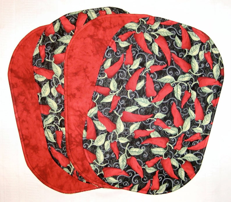 Puede incluir: Juego de cuatro manteles individuales ovalados de color rojo y negro con un patr&oacute;n de chiles. Los manteles individuales tienen un borde rojo y un fondo negro con chiles rojos y hojas verdes.