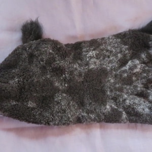 Puede incluir: Un juguete de peluche gris oscuro y blanco con una pequeña cola y una etiqueta sujeta por un anillo de metal. El juguete tiene un aspecto texturizado y peludo y está sobre una superficie de color púrpura claro.
