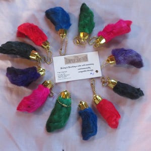 May include: A collection of 12 colorful faux rabbit feet keychains with gold rings and chains. The keychains are arranged in a circle on a white surface. The text "King & Trading, a cabin with amazing, environmentally compatible things" is visible on a card in the center of the circle.