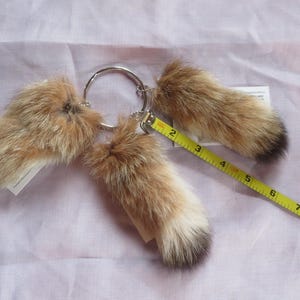May include: A set of three fluffy, light brown and black tipped fur tails attached to a silver metal ring. A yellow measuring tape is next to the tails. The tails are approximately 4 inches long.