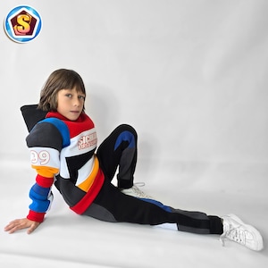 Può includere: Tuta colorata per bambini, composta da felpa con cappuccio e pantaloni abbinati. La felpa è rossa, bianca, blu e gialla con la scritta "SICHKA FIRST FASHION" e il numero "39". I pantaloni sono neri con dettagli grigi e blu. Le scarpe da ginnastica bianche completano l'outfit.