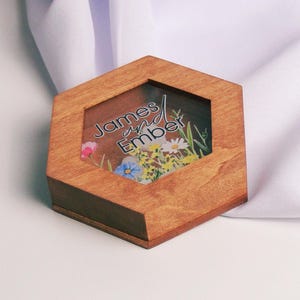 May include: A wooden hexagonal ring box with a clear lid. The lid features a floral design and the text "James and Ember".