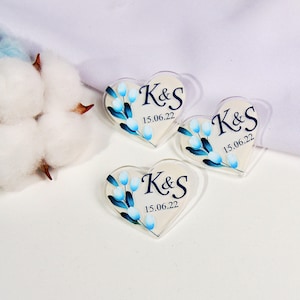 May include: Three heart-shaped acrylic favors with floral designs and the initials "K&S" and the date "15.06.22" in navy blue. The favors have a clear border and are set against a white background. Cotton boll and white fabric are in the background.
