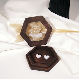 Puede incluir: Una caja de anillos hexagonal de madera con tapa de cristal. La tapa tiene los nombres "Jason & Rachel" grabados con un diseño de pluma. La caja está abierta y hay dos almohadones en forma de corazón blanco en el interior.