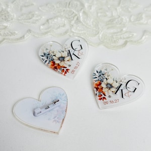 May include: Three clear acrylic heart-shaped pins with floral designs and the initials "GK" and the date "09.16.27". One pin has a pin back attached. The floral design includes white, orange, and blue flowers and leaves.