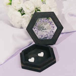Puede incluir: Una caja de anillos hexagonal de madera con una tapa transparente. La tapa tiene un diseño floral con flores moradas y el texto "August & Donavan 2023". La caja está abierta y tiene dos almohadas blancas en forma de corazón en el interior.