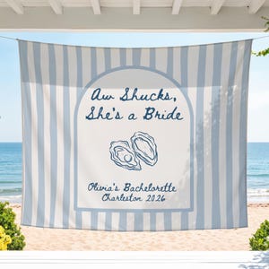 Może przedstawiać: Niebiesko-biały ręcznik plażowy w paski z napisem "Aw Shucks, She's a Bride" i "Olivia's Bachelorette Charleston 2026". Ręcznik przedstawia ilustrację dwóch ostryg.