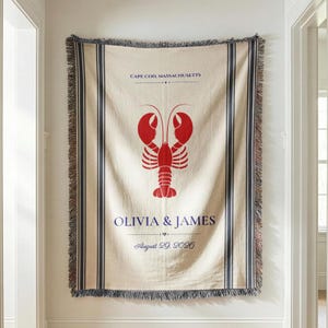 May include: A cream-colored woven throw blanket featuring a large red lobster graphic in the center. The text reads CAPE COD, MASSACHUSETTS, OLIVIA & JAMES, and August 29, 2026. The blanket has dark blue vertical stripes and fringed edges on all sides.