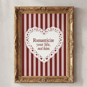 Könnte beinhalten: Gerahmter Druck mit einem verzierten goldenen Rahmen. Der Hintergrund hat vertikale rote und rosa Streifen. Eine herzförmige weiße Spitze mit dem Text "Romanticize your life, not him." ist auf dem Druck zentriert.