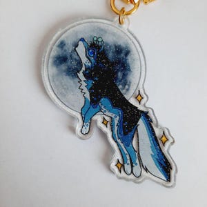 Könnte beinhalten: Ein klarer Acryl-Schlüsselanhänger mit einem heulenden Wolf vor einem Vollmond. Der Wolf ist blau und schwarz mit Glitzer. Der Mond ist hellblau mit Glitzer, und es gibt goldene Sternakzente.