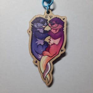 Könnte beinhalten: Holzschlüsselanhänger mit blauem Ring, der zwei Cartoon-Otter in einer Umarmung zeigt. Einer der Otter ist lila mit Sternenmuster, der andere ist rosa und rot. Der Schlüsselanhänger hat einen Regenbogen-Schwanz.