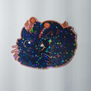 Könnte beinhalten: Ein holografischer Aufkleber mit einer Cartoon-Katze mit dunkelblauem Körper und bunten Sternakzenten. Die Katze hat große, ausdrucksstarke Augen und ist kupferfarben umrandet. Der Aufkleber hat ein glitzerndes, reflektierendes Finish.