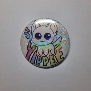 Könnte beinhalten: Ein runder Button mit holografischem Hintergrund und einer Cartoon-Kreatur-Illustration. Die Kreatur ist weiß mit großen Augen und einem kleinen Mund. Das Wort "YIPPEE" steht in bunten Buchstaben darunter.