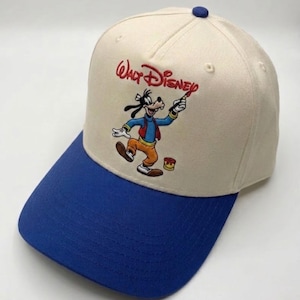 Op de afbeelding: Een baseballpet met een crèmekleurige kroon en een blauwe rand. De pet heeft een geborduurd beeld van Goofy van Disney, met een penseel, met de woorden "Walt Disney" erboven. De pet heeft een blauwe knop bovenop.