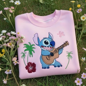 May include: A light pink sweatshirt featuring an embroidered cartoon character playing a ukulele. The design includes palm trees, flowers, and the character in blue. The sweatshirt is made of a soft material.