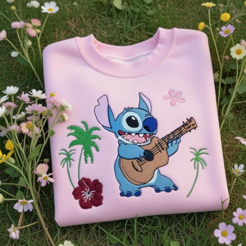 May include: A light pink sweatshirt featuring an embroidered cartoon character playing a ukulele. The design includes palm trees, flowers, and the character in blue. The sweatshirt is made of a soft material.