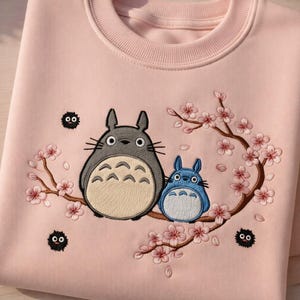 May include: A pale pink sweatshirt with an embroidered design of two Totoro characters and a flowering branch with pink blossoms. The design includes small black soot sprites. The sweatshirt has a crew neck.