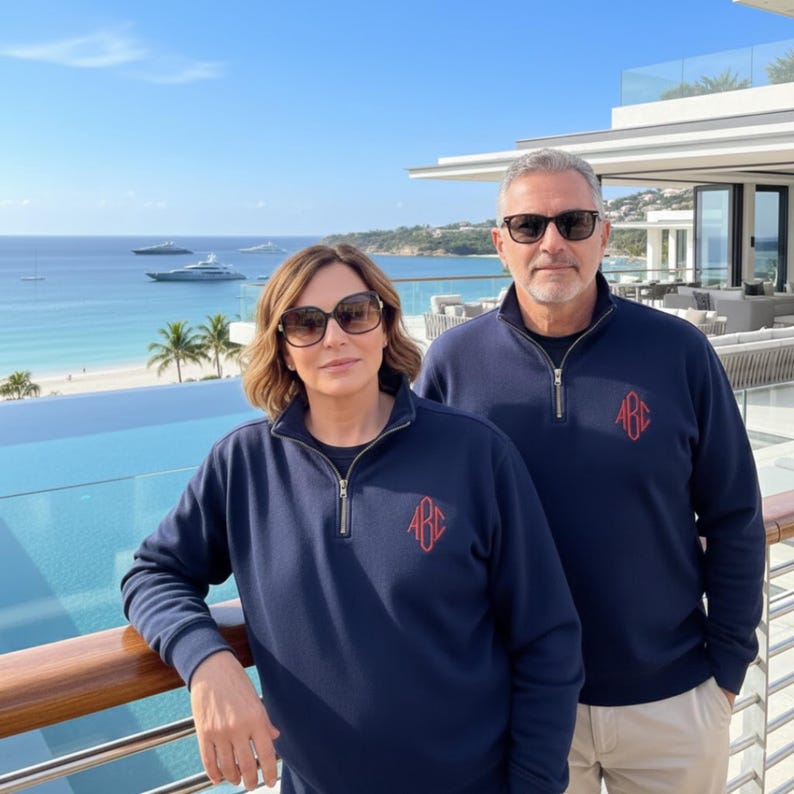 May include: Two navy blue quarter-zip pullovers with red monogram embroidery. The pullovers have a stand-up collar and a zipper closure. The background shows a scenic view of the ocean and a luxurious outdoor setting.