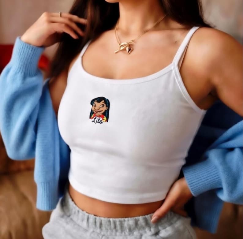 May include: White crop top with thin straps featuring a colorful embroidered image of Lilo from the Disney movie Lilo & Stitch. The top is worn with a light blue cardigan and gray sweatpants. A gold necklace is also visible.
