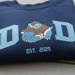 May include: Navy blue sweatshirt with the word "DAD" in light blue block letters. A cartoon turtle character is embroidered in the center. The text "EST. 2025" is below the turtle. The sweatshirt is folded.