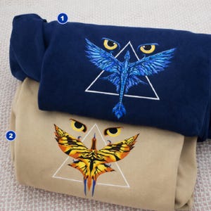 May include: Two sweatshirts, one navy blue and one tan, are stacked. Each features an embroidered design of a stylized bird within a white triangle. The navy sweatshirt has a blue bird design, while the tan one has a yellow and orange bird.