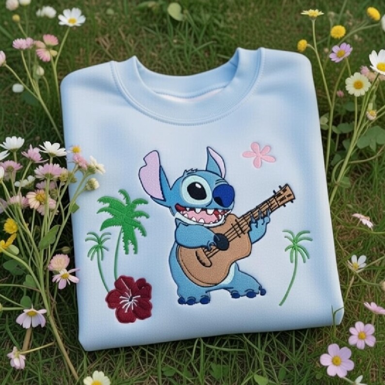 May include: A light blue sweatshirt featuring an embroidered cartoon character playing a guitar. The design includes palm trees and flowers in green, pink, and red. The sweatshirt is displayed on a bed of green grass with flowers.