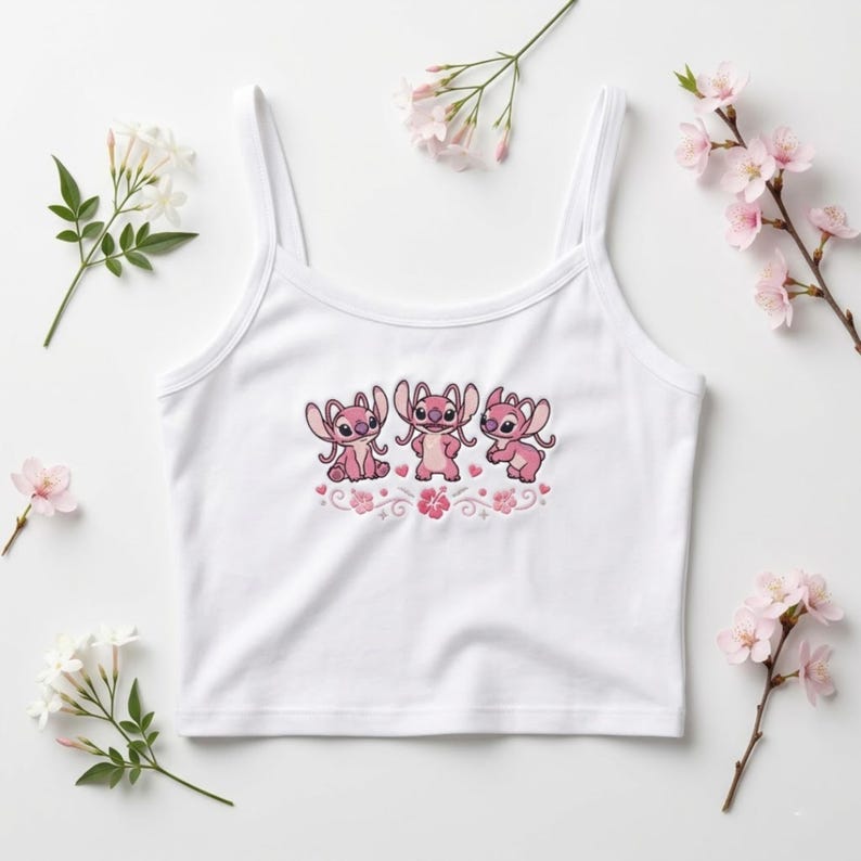 May include: White tank top with spaghetti straps. The front features an embroidered design of three pink cartoon characters with large ears and a floral border. The design is in shades of pink and red.