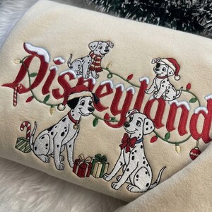 May include: A beige sweatshirt featuring embroidered Disneyland text in red with snow accents. The design includes four Dalmatian puppies, some wearing Santa hats or scarves, surrounded by a string of Christmas lights and colorful gift boxes.