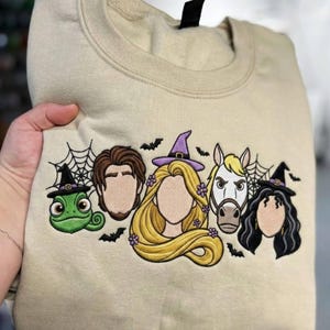 Könnte beinhalten: Ein beigefarbenes Sweatshirt mit Rundhalsausschnitt und einer Stickerei von fünf Zeichentrickfiguren. Das Motiv zeigt ein grünes Chamäleon, einen Mann mit braunen Haaren, eine blonde Frau mit violettem Hexenhut, ein weißes Pferd und eine Frau mit schwarzem Haar.