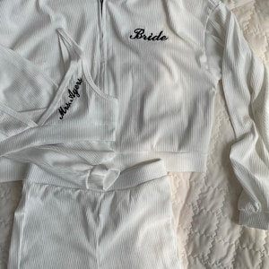 May include: A white ribbed three-piece lounge set consisting of a zip-up hoodie, a bralette, and shorts. The hoodie features the word Bride embroidered in black. The bralette displays the text Mrs. Ayers, and the shorts feature the word Bride.