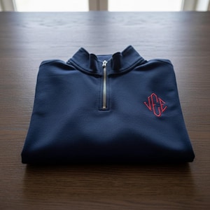 May include: Navy blue quarter-zip sweatshirt with a red embroidered monogram. The folded sweatshirt features a stand-up collar and a silver zipper. The monogram is in a stylized font. The sweatshirt is made of a soft material.