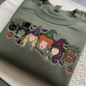 May include: An olive green crewneck sweatshirt featuring detailed embroidery of three witch characters with distinct hair colors, wearing pointed hats. The design includes a black cat, spiderwebs, small pumpkins, a broom, and a spellbook.